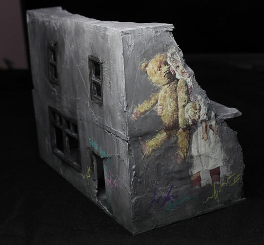 A corner view of a grey box modern building, with a large mural of a child with a teddy bear on the side. The mural is partially destroyed, along with the wall.